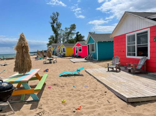 Colorful beach cabins line a sandy shore under a bright blue sky, with chairs and beach toys scattered around.