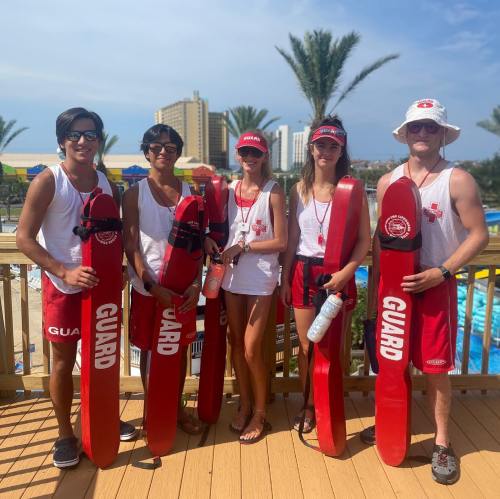 Five lifeguards in red uniforms stand together by a pool, holding rescue boards, with palm trees and buildings in the background.