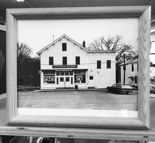 Black and white photo of a vintage general store with a car parked outside, framed in wood.