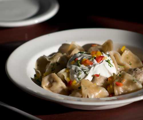 A plate of dumplings topped with sour cream and colorful diced peppers, served on a wooden table.