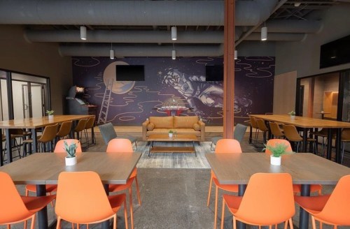 A modern dining area with orange chairs, wooden tables, and a mural of a figure on a ladder against a dark wall.