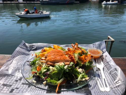 A plate of salad with salmon, served on a table by the water, with boats in the background.