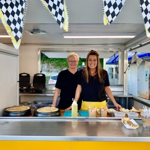 Two smiling vendors stand behind a food cart, preparing crepes, with checkered bunting overhead.