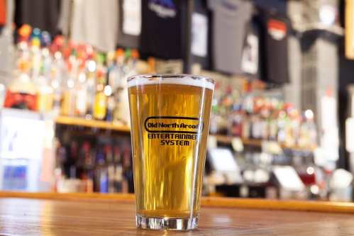 A pint of beer sits on a wooden bar, with a colorful backdrop of bottles and merchandise in a lively setting.