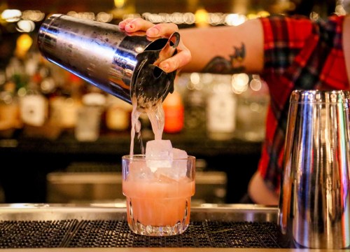 A bartender pours a cocktail from a shaker into a glass with ice, surrounded by blurred bottles in the background.