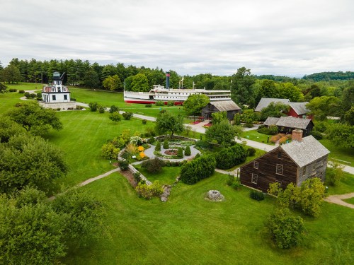 Aerial view of a historic site featuring a ship, gardens, and several rustic buildings surrounded by greenery.