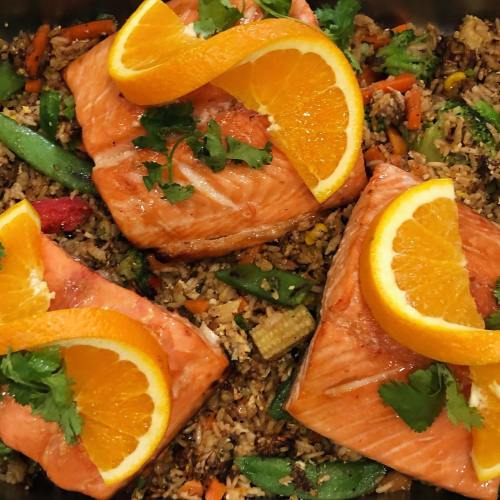 Two salmon fillets topped with orange slices, served on a bed of colorful fried rice with vegetables.