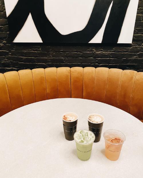 Four drinks on a round table with a brown leather bench and a black and white abstract painting in the background.