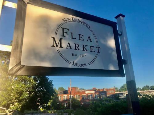 Sign for New Brunswick Flea Market, established in 2017, with "Indoor" noted below. Clear blue sky in the background.