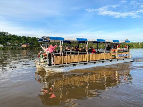 A tour boat with passengers cruising on a river, surrounded by greenery and a clear blue sky.