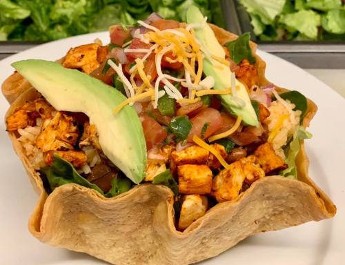 A colorful taco salad in a crispy tortilla bowl, filled with chicken, rice, lettuce, tomatoes, cheese, and avocado.