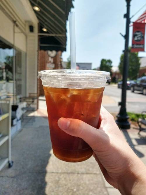 A hand holding a clear cup of iced coffee with a straw, set against a sunny street scene.