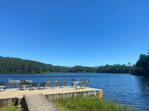 A serene lake view with a wooden dock and chairs, surrounded by lush greenery and clear blue skies.