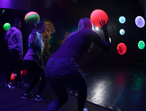 Three people in a dark room are playing with glowing balls, with colorful planets projected on the wall behind them.