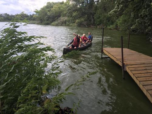 Three people in a canoe approach a wooden dock on a calm, green river surrounded by lush trees.