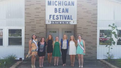 Group of eight women in colorful outfits posing in front of the Michigan Bean Festival entrance sign.