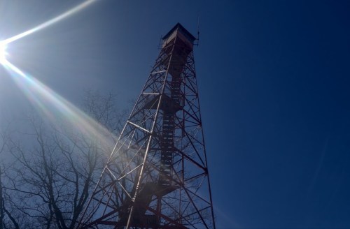 A tall red metal tower against a clear blue sky, with sunlight creating a lens flare effect.