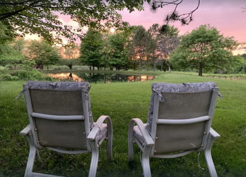 Two white lawn chairs face a serene pond surrounded by lush greenery and trees, with a colorful sunset in the background.