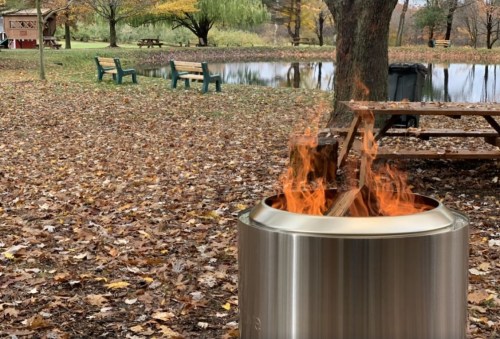 A fire pit with flames burning, surrounded by autumn leaves and a peaceful park setting with benches and a pond.