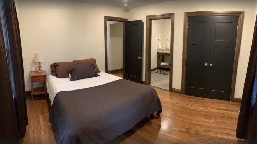 A cozy bedroom with a bed, nightstand, and two doors leading to other rooms, featuring wooden floors and dark trim.