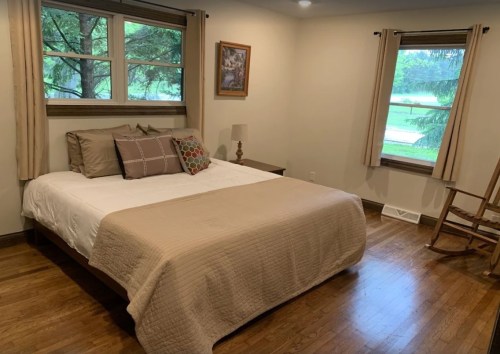 Cozy bedroom with a bed, decorative pillows, a rocking chair, and large windows overlooking greenery.