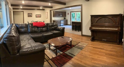 Cozy living room with a leather sectional, coffee table, and a vintage piano, featuring warm wood floors and natural light.