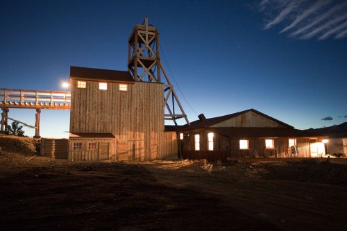 A wooden mining structure illuminated at night, with a tall tower and a building beside it under a starry sky.