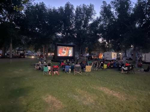A crowd sits on lawn chairs watching a movie on a large screen in a park at dusk, surrounded by trees and RVs.