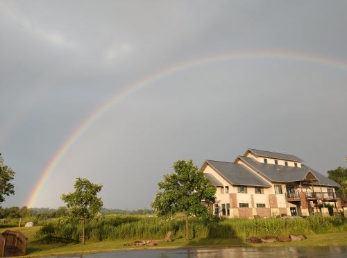 A rainbow arcs over a modern building surrounded by greenery and a calm water surface.