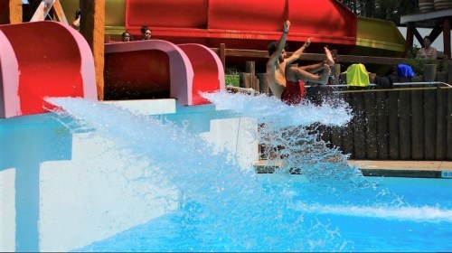 Two people splash into a pool from colorful water slides, with onlookers in the background enjoying the water park.