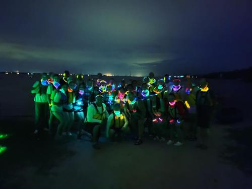 A large group of people wearing glowing accessories poses together at night near a body of water.