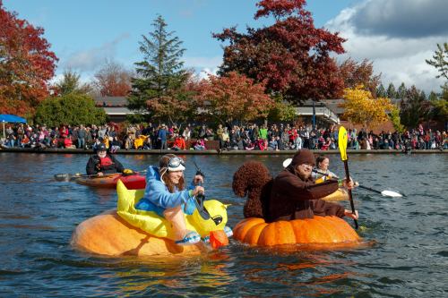 Two people in costume float on giant inflatable pumpkins and a duck in a lively autumn scene with a crowd in the background.