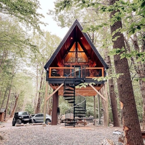 A modern A-frame cabin with a spiral staircase, surrounded by trees and parked cars in a serene forest setting.