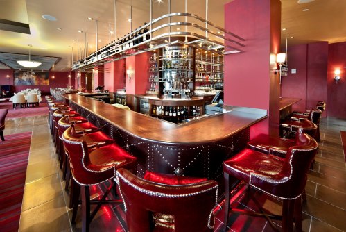 A stylish bar with a curved wooden counter, red leather stools, and a backdrop of shelves filled with bottles.