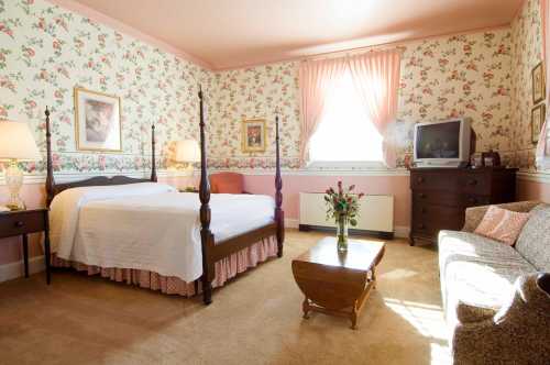 Cozy bedroom with floral wallpaper, a four-poster bed, a TV, and a small seating area with a coffee table.