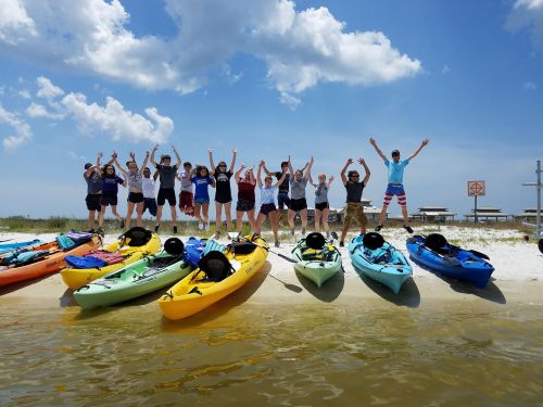 A group of people joyfully jumping on a beach near colorful kayaks under a bright blue sky.