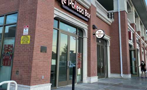 Exterior view of The Pollo Bar, featuring brick walls and large glass doors, with a person walking by.