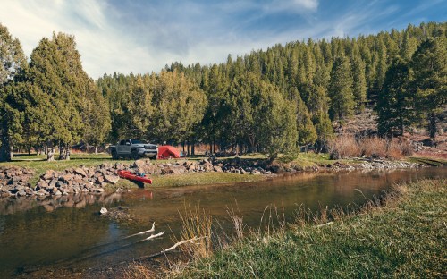 A serene campsite by a river, featuring a tent, a kayak, and surrounded by lush green trees and rocky terrain.