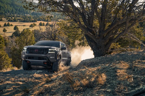 A rugged truck drives off-road, kicking up dust near a large tree in a scenic, mountainous landscape.
