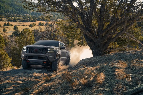 A rugged truck driving off-road through a dusty landscape with trees and mountains in the background.