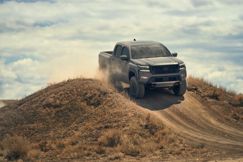 A rugged truck driving over a dirt mound, kicking up dust against a backdrop of cloudy skies.