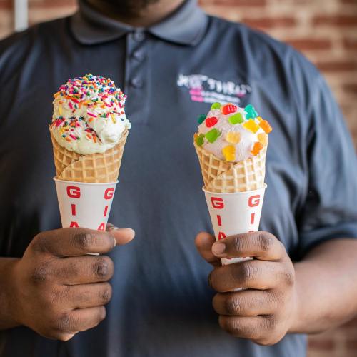 A person holds two ice cream cones, one topped with sprinkles and the other with colorful candy pieces.