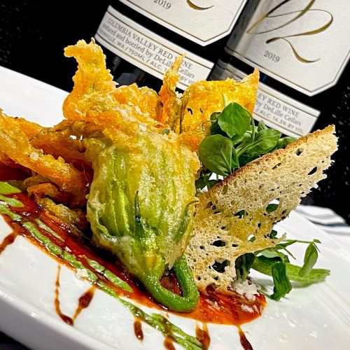 A gourmet dish featuring stuffed zucchini blossoms, greens, and a decorative cheese crisp on a plate with sauce drizzles.