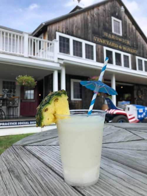A refreshing cocktail with a pineapple slice and umbrella, set on a wooden table in front of a vineyard building.