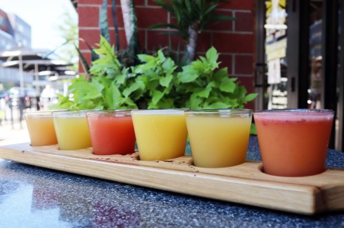 A wooden flight board holds six colorful drinks, ranging from yellow to red, set against a backdrop of greenery.