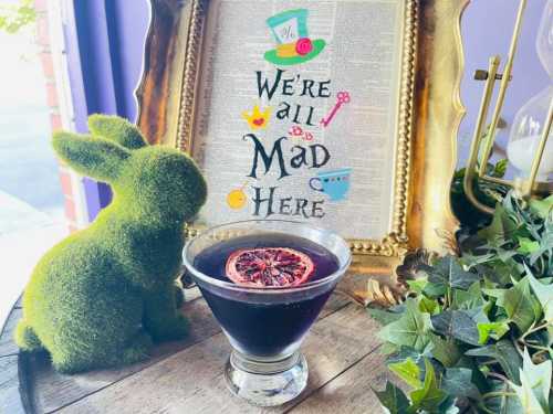 A green bunny decoration beside a cocktail with a dried orange slice, framed by a whimsical "We're all Mad Here" sign.