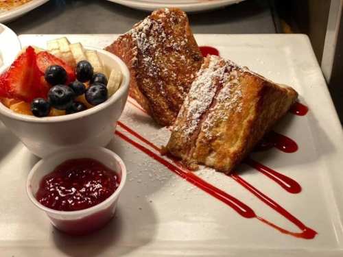 Plate of French toast dusted with powdered sugar, served with fresh fruit and a side of raspberry sauce.