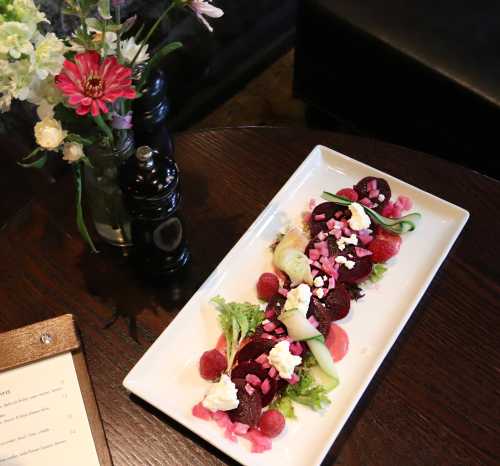 A colorful salad with beets, greens, and toppings, served on a white plate, next to a vase of flowers.