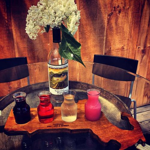 A wooden tray holds four small bottles of colorful liquids, with a wine bottle and white flowers in the background.