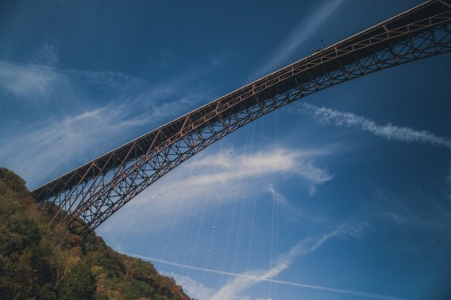 A large metal bridge spans across a blue sky with wispy clouds, surrounded by green hills.
